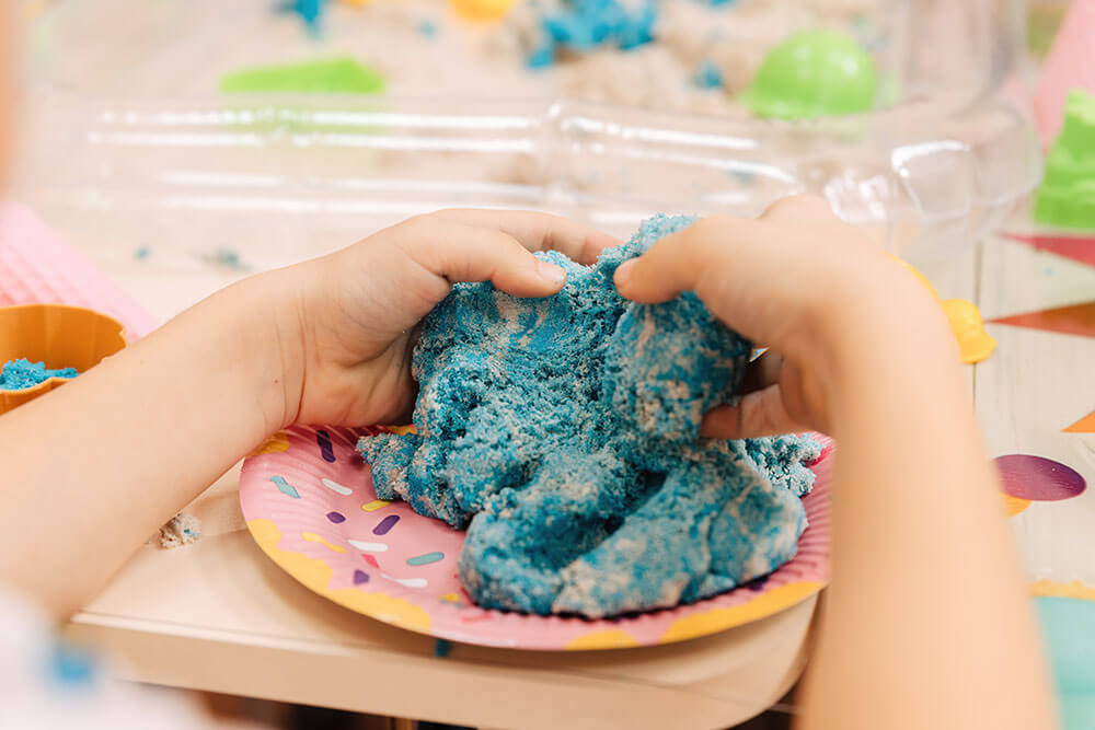 child hands shaping blue kinetic sand on a colorful plate