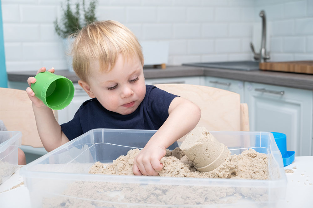 Child playing with kinetic sand using colorful cups in a kitchen setting