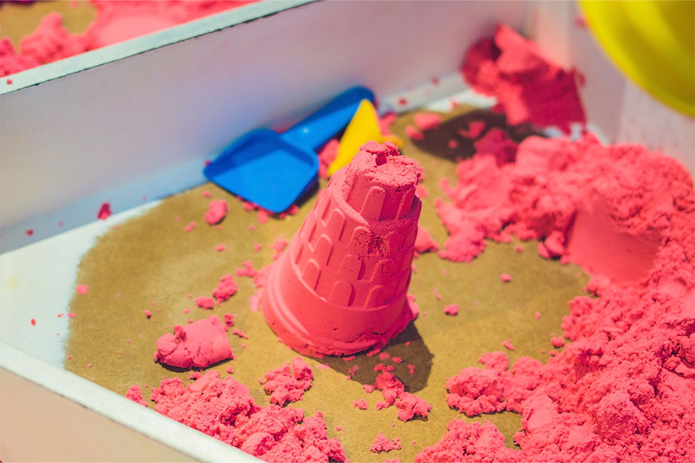 Pink kinetic sand castle mold with scattered sand pieces and blue/yellow tools nearby